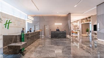 A modern hotel lobby with a reception desk, a waiting area, and a staircase leading to upper floors.
