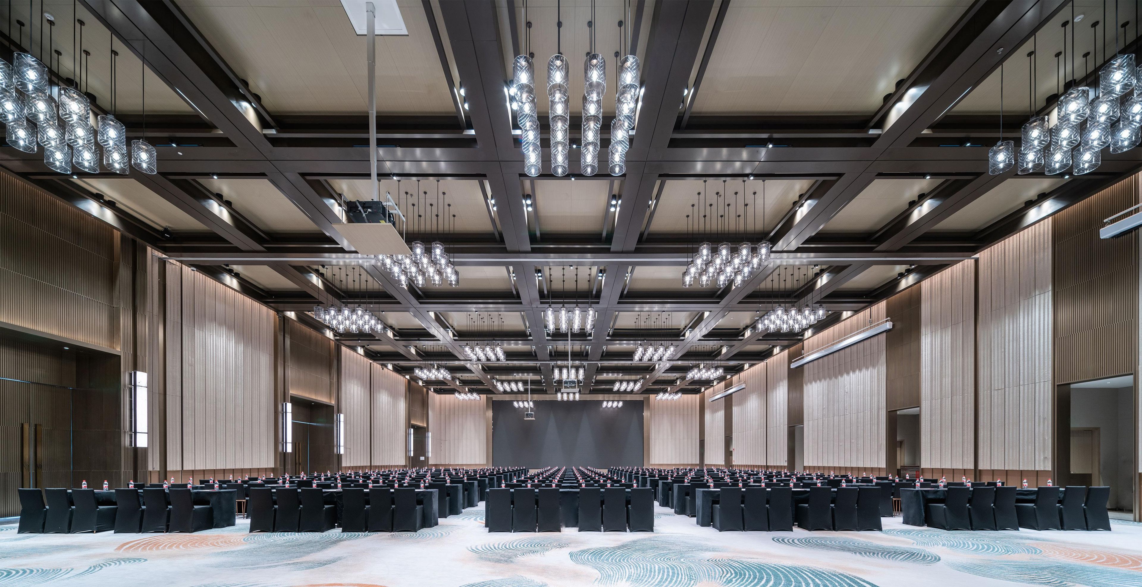 A large conference room with rows of chairs, a high ceiling with hanging lights, and a patterned carpet.