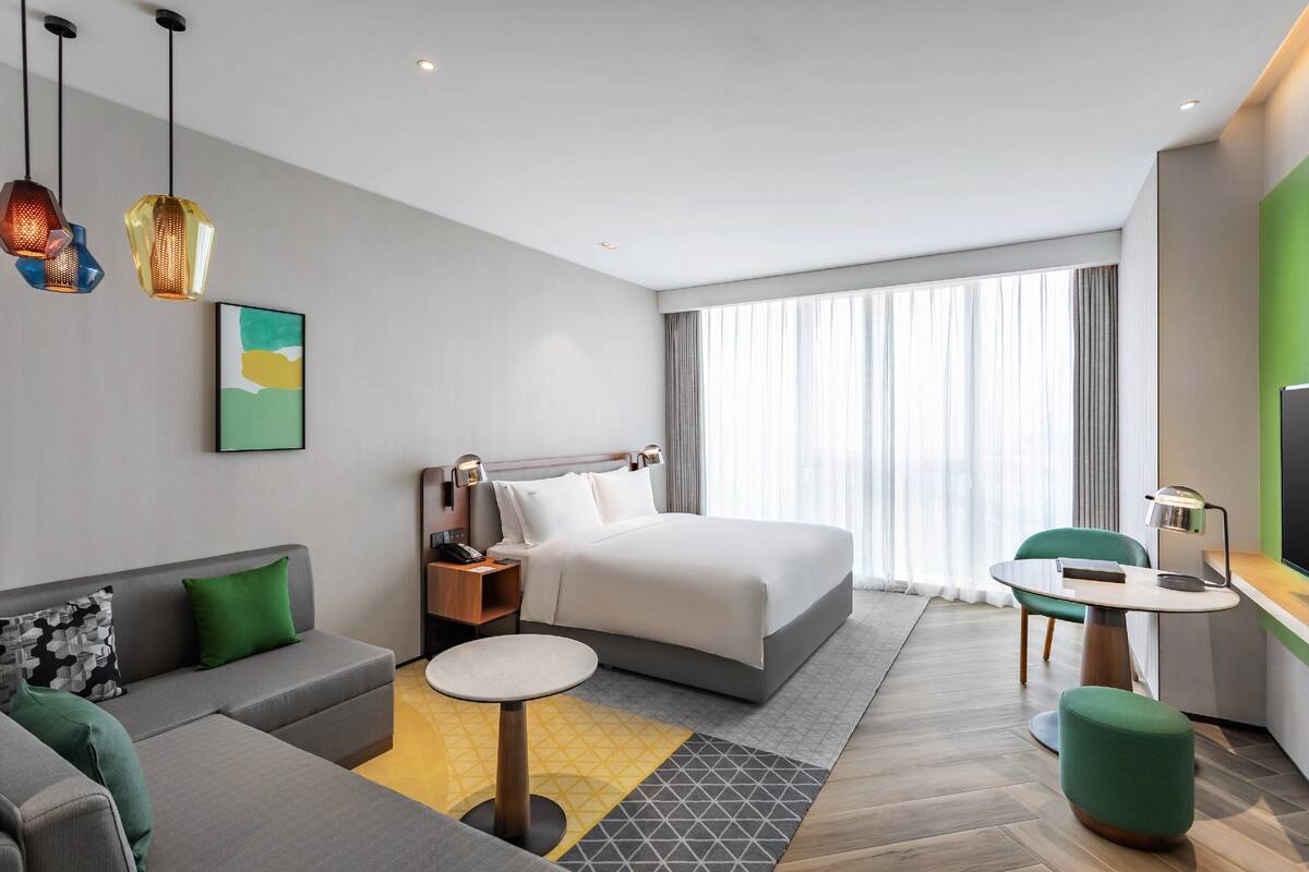 A modern hotel room with a bed, sofa, desk, and a large window with curtains.