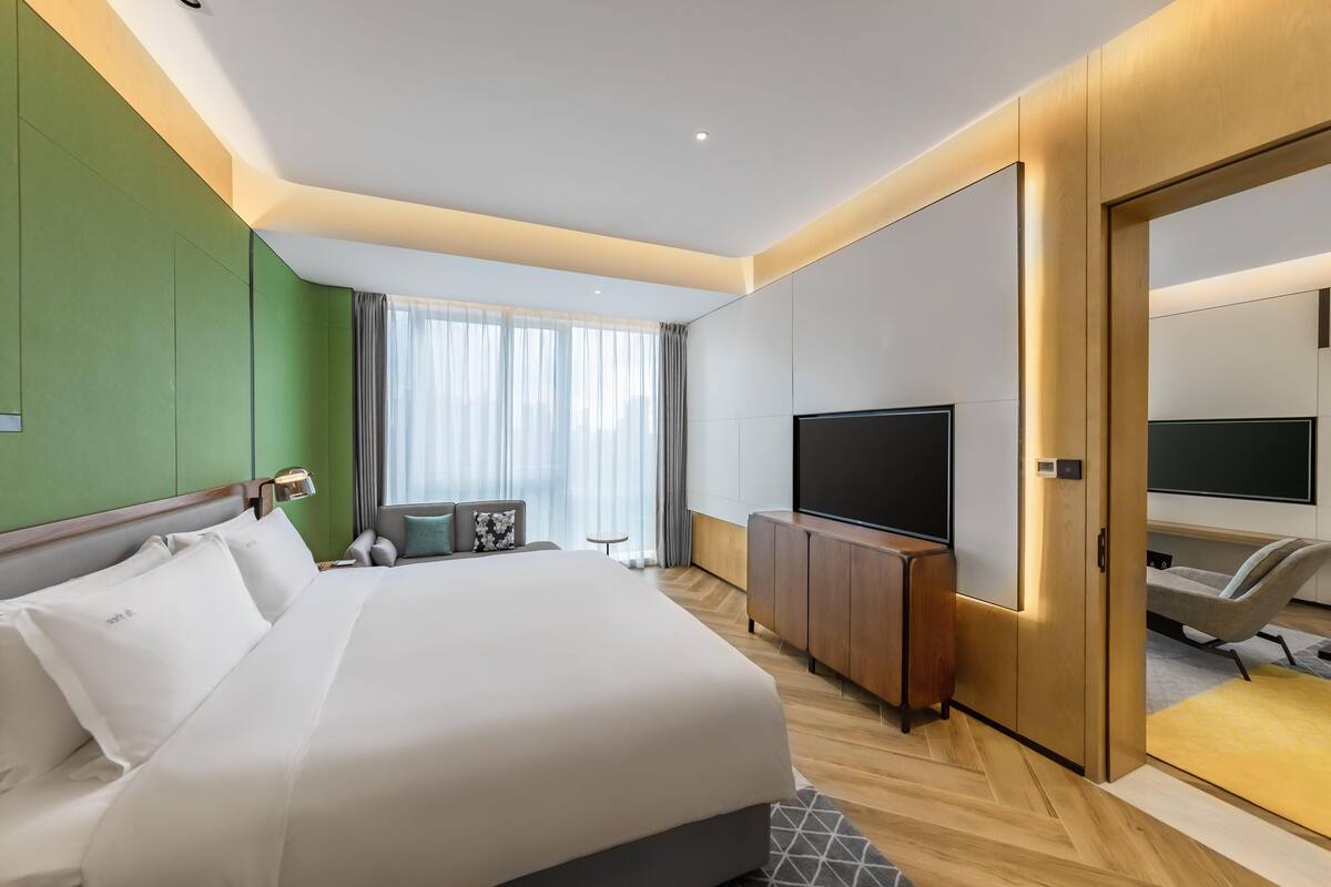 A modern hotel room with a large bed, a TV, a seating area, and a green accent wall.
