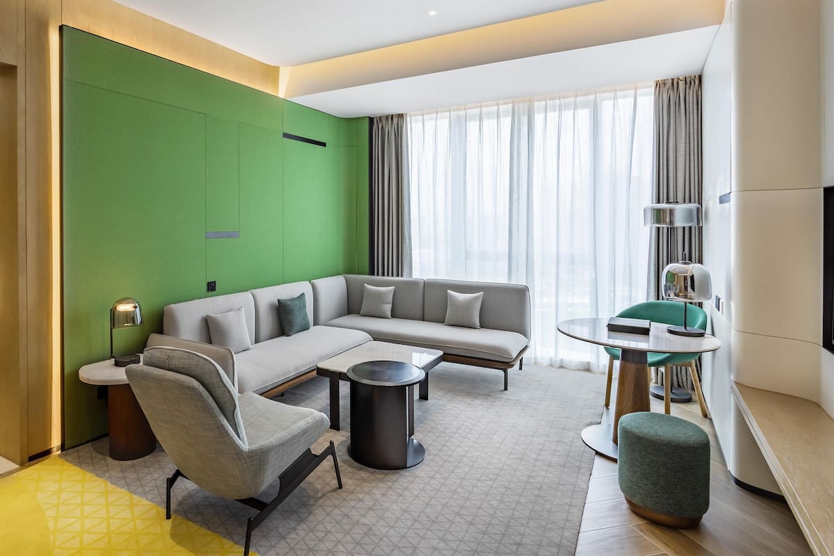 A modern hotel room with a green accent wall, a sofa, a coffee table, a chair, a TV, and a bed.