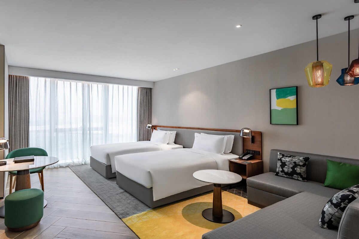 Hotelkamer met twee bedden, een bank, een bureau met stoel, een televisie en een groot raam met gordijnen.