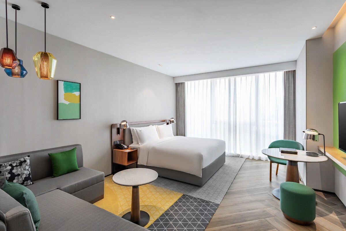 Een moderne hotelkamer met een bed, een bank, een bureau en een groot raam met gordijnen.