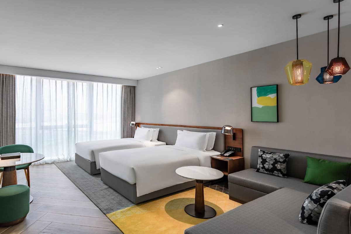 A hotel room with two beds, a sofa, a desk with a chair, a TV, and a large window with curtains.