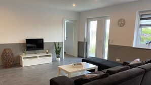 Uma sala de estar moderna com um sofá grande e escuro, uma mesa de centro branca e uma TV de tela plana sobre um móvel branco. Há uma planta em vaso e um vaso decorativo no móvel.
