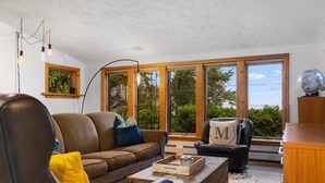A modern living room with a brown sofa, a black leather chair, and a wooden coffee table. There are large windows with a view of trees and a clear sky. - 2 Bedroom Carriage House with view of Suttons Bay (Suttons Bay)