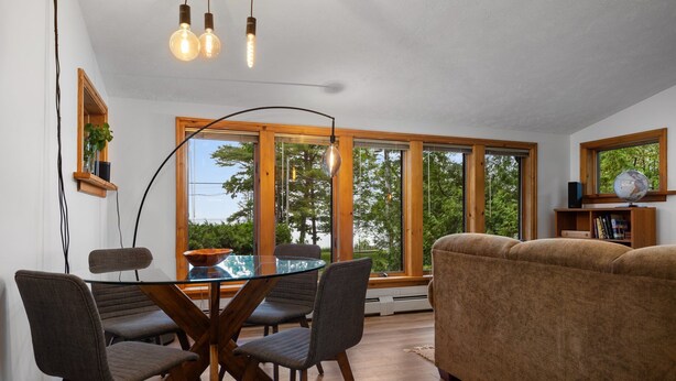 A modern living room with a glass dining table, four chairs, a brown sofa, and large windows with a view of trees. - 2 Bedroom Carriage House with view of Suttons Bay (Suttons Bay)
