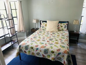 A bedroom with a bed, two bedside tables, a nightstand, a window with curtains, a chair, and a blue rug. - 2,2 Stilted House on private peninsula. Renovated June '23.  (Homosassa)