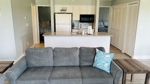 Ein modernes Wohnzimmer mit grauem Sofa, einem Holztisch und einer Küchenzeile mit weißen Schränken und einer schwarzen Mikrowelle.