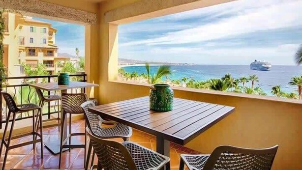 Ein Balkon mit Tisch und Stühlen, mit Blick auf einen Strand und den Ozean, wobei sich in der Ferne ein Kreuzfahrtschiff befindet.