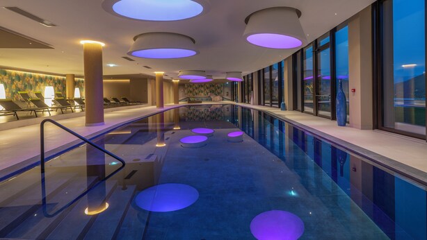 A modern indoor pool with circular lighting fixtures, a glass balustrade, and a view of the cityscape through large windows.