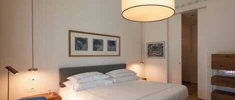 A bedroom with a bed, bedside tables, a chair, a hanging light fixture, and framed artwork on the wall.
