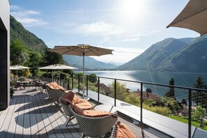 A deck with outdoor furniture and umbrellas overlooking a lake with mountains in the background. - Aria Retreat & SPA (Porlezza)