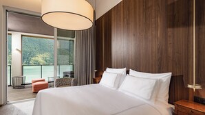 A hotel room with a large bed, wooden panel walls, a balcony with a view of a lake, and a modern chandelier.