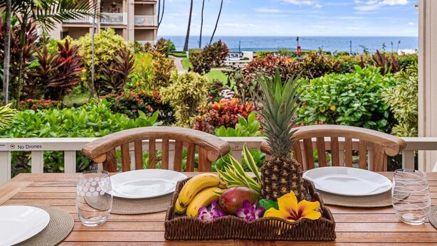 A dining table with a fruit basket, two chairs, and a view of the ocean and hotel buildings.
