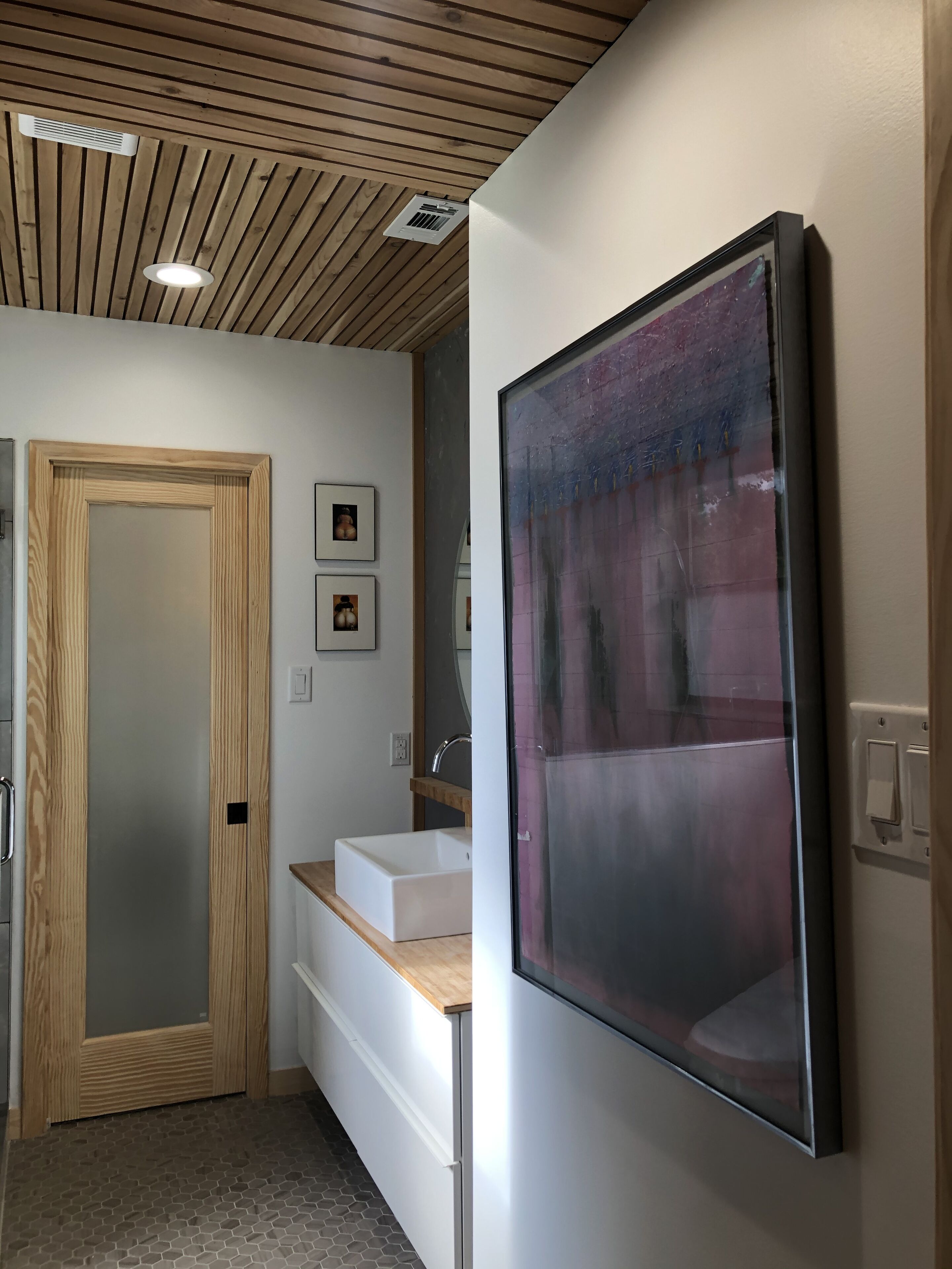 A modern bathroom with a wooden door, a white sink, and a large abstract painting on the wall.
