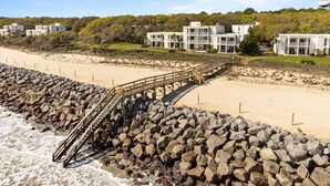 Et kysthotel med en træbro, der fører til en sandstrand, omgivet af klippeafsatser og frodig vegetation.