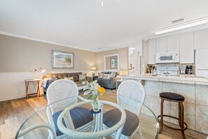 A modern living room with a glass dining table, two white wicker chairs, a sofa, and a kitchen area with white cabinets and a microwave.