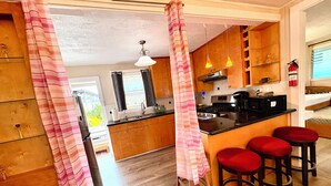 A kitchen with wooden cabinets, a black countertop, a stove, and a sink. There are four red stools and a striped curtain.