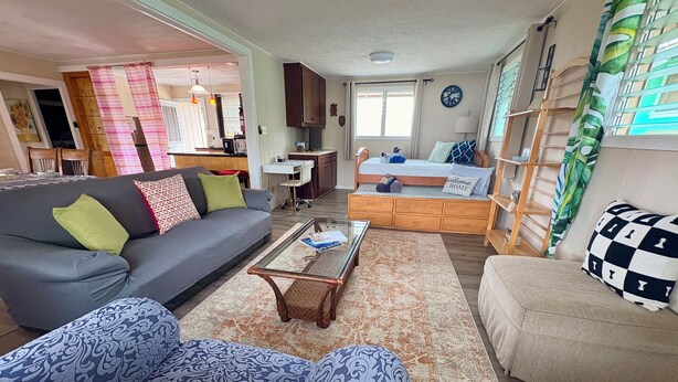A living room with a sofa, armchairs, a coffee table, and a bed with a headboard. There is a dining area in the background with a table and chairs.
