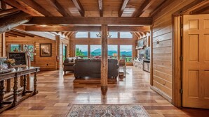 A spacious log cabin interior with a wooden ceiling, a fireplace, a sofa, and a view of the mountains.