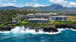 A coastal hotel with multiple buildings, a rocky shoreline, and a mountainous backdrop. - Expansive Views with new split system AC! (Koloa)