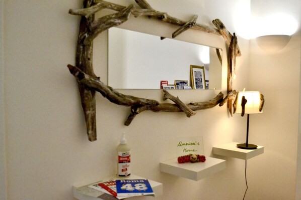 A room with a mirror framed by a natural branch, a bedside table with a lamp, and a small shelf with a sign reading "America's Home". - Amerina's Home (Roma)