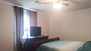 Un dormitorio con cama, televisor sobre un mueble, ventilador de techo y ventana con cortinas.