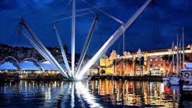 Un hotel moderno frente al agua, con una estructura central destacada y vigas que se extienden, un puerto deportivo con barcos y un paisaje urbano al fondo.