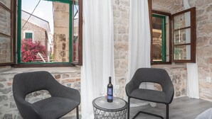 Una habitación con paredes de piedra, dos sillas grises, una mesita redonda con una botella y dos vasos, y una ventana con contraventanas verdes.
