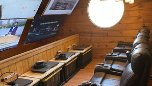 A wooden-paneled room with a TV mounted on the wall displaying a movie. There are leather recliners and a wooden counter with electronic equipment.