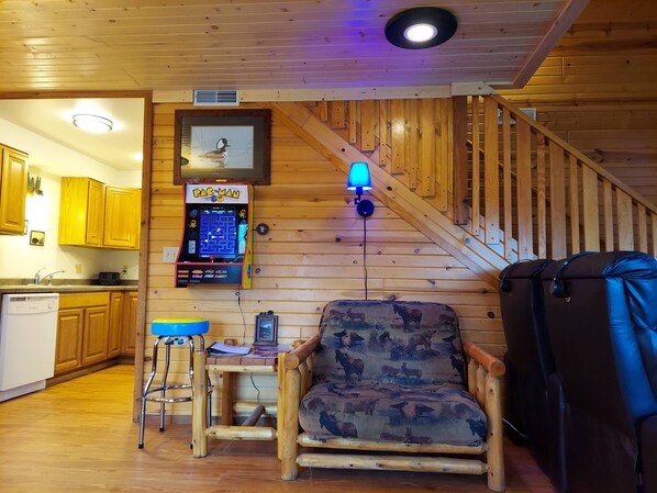 A cozy cabin interior with a wooden staircase, a dining area with a table and chairs, and a Pac-Man arcade game.