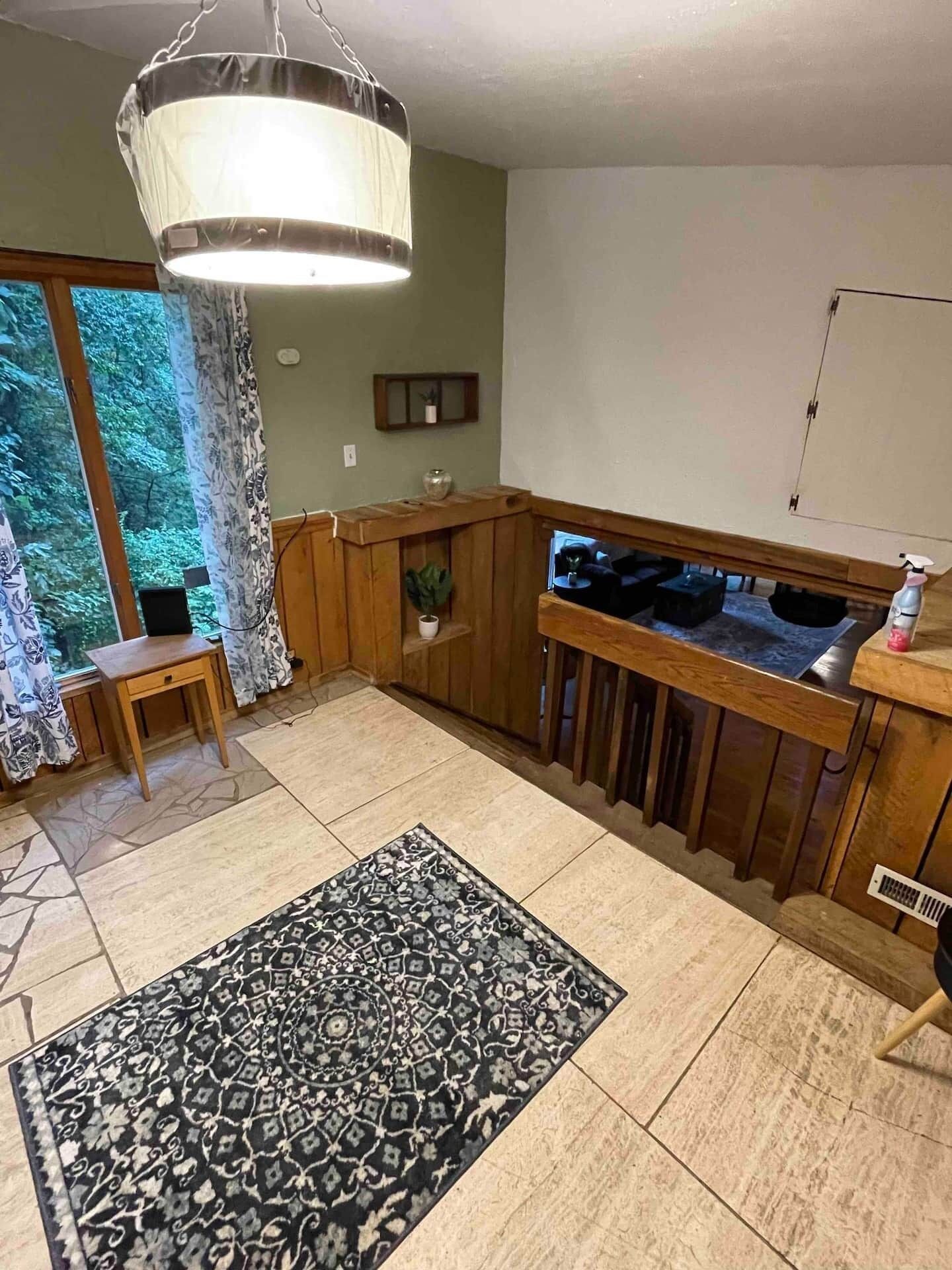 A room with a wooden desk, a chair, a patterned rug, a hanging light fixture, and a view through a window.