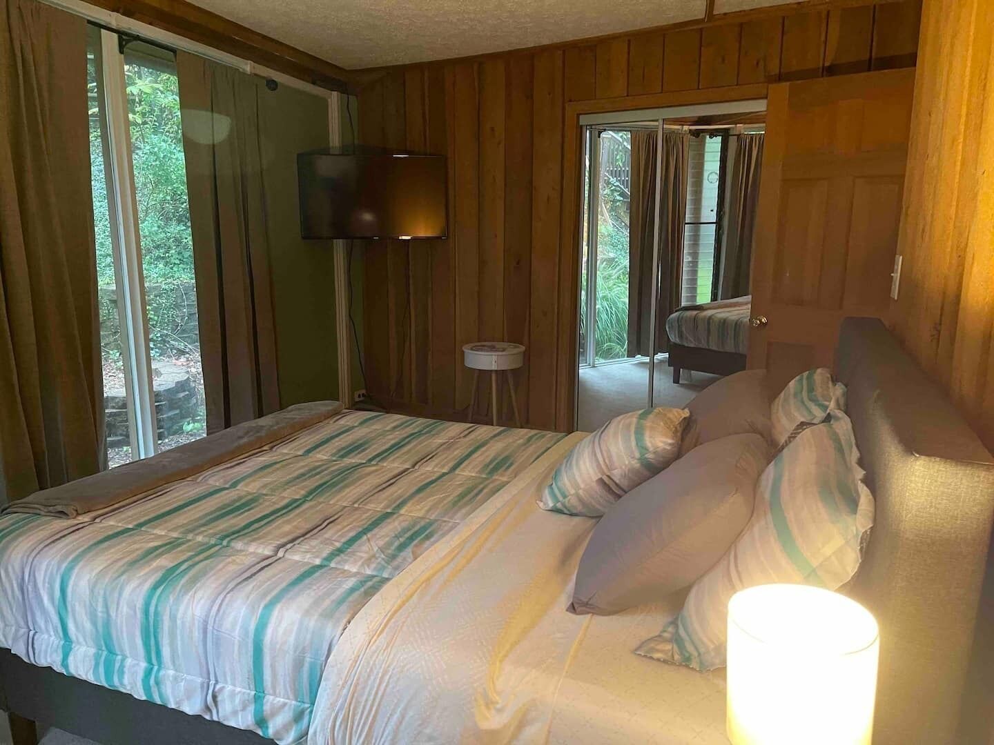 A bedroom with a bed, a bedside lamp, a television on the wall, and a view of the outdoors through a sliding door.