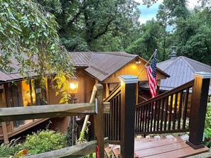A wooden cabin with a porch and a staircase leading to a deck, surrounded by lush greenery.