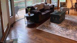 A spacious living room with a wooden floor, a dark-colored sofa, a small round table, and a decorative chest. Large windows provide a view of the outdoors. - TreeTops in Town Private & Peaceful (Charleston)