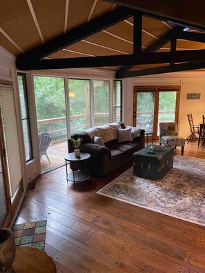 A spacious living room with a wooden floor, a dark-colored sofa, a small round table, and a decorative chest. Large windows provide a view of the outdoors. - TreeTops in Town Private & Peaceful (Charleston)