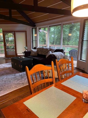 A cozy living room with a wooden table, chairs, a sofa, and a large window offering a view of trees.