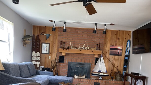 Un salón con chimenea, piso de madera, ventilador de techo y televisor montado.