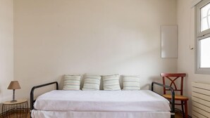 Una camera da letto con un letto, quattro cuscini a righe, un piccolo tavolino con una lampada, una sedia e una finestra.
