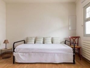 A bedroom with a bed, four striped pillows, a small round table with a lamp, a chair, and a window. - Mogador by Interhome (Arcachon)