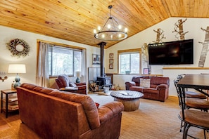 Ein gemütliches Wohnzimmer mit Ledersofas, Holzbalkendecke, Kamin, Fernseher und großen Fenstern.
