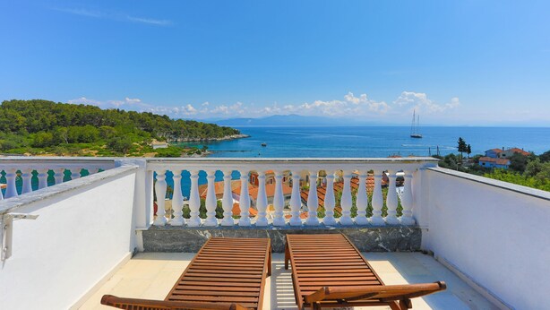 A balcony with two wooden chairs facing a scenic view of a coastal area with buildings and a clear blue sky.