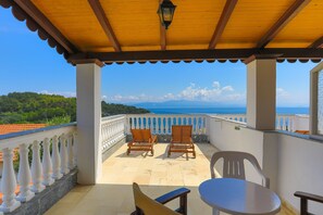 A covered balcony with two wooden chairs and a table, overlooking a scenic view of the sea and mountains.