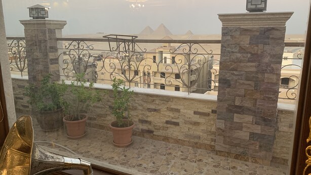 Un balcone con piante in vaso, una ringhiera in metallo e una vista di edifici e piramidi all'orizzonte.