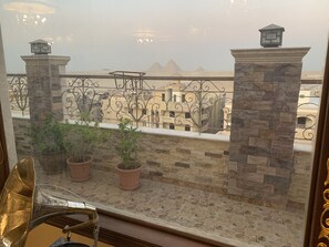 A balcony with potted plants, a metal railing, and a view of buildings and pyramids in the distance.