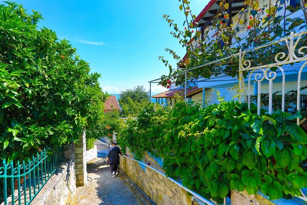 A narrow alleyway with a stone wall and metal railing on one side, leading to a building with a blue roof and a person walking away.