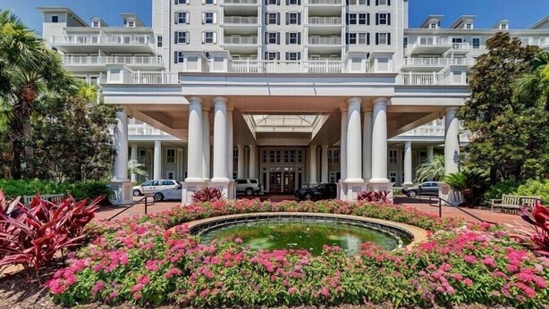 A multi-story hotel with a central entrance flanked by columns, surrounded by a landscaped garden with a circular pond and pink flowers. - Enjoy This Elegant Coastal Condo:, King-Suite,Full kitchen,Pool, Hot Tub,Balcony (Miramar Beach)