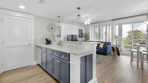 A modern kitchen with a white island, dark cabinets, and a blue sofa in the living area. - Enjoy This Elegant Coastal Condo:, King-Suite,Full kitchen,Pool, Hot Tub,Balcony (Miramar Beach)