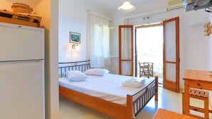 Un dormitorio con una cama, una mesa de madera, una silla, un refrigerador y una puerta que da a un balcón.